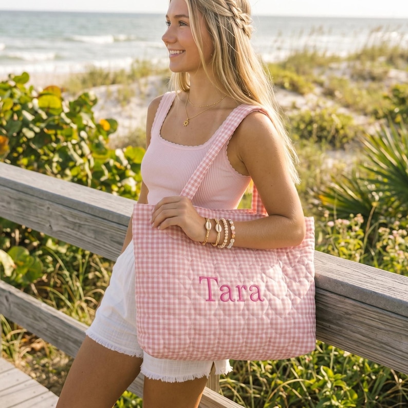 May include: A quilted tote bag featuring a light pink and white gingham pattern with the name Tara embroidered in pink thread on the front. The bag has two matching gingham shoulder straps and a rectangular shape with a soft, padded texture.