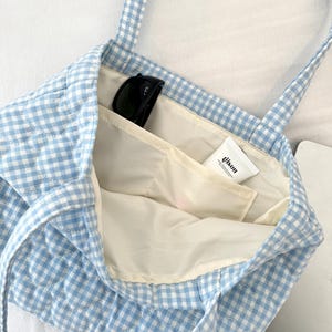 May include: A light blue and white gingham quilted tote bag with two shoulder straps. The interior is lined with cream fabric and features an open pocket containing a pair of black sunglasses and a white tube labeled with the text gison.