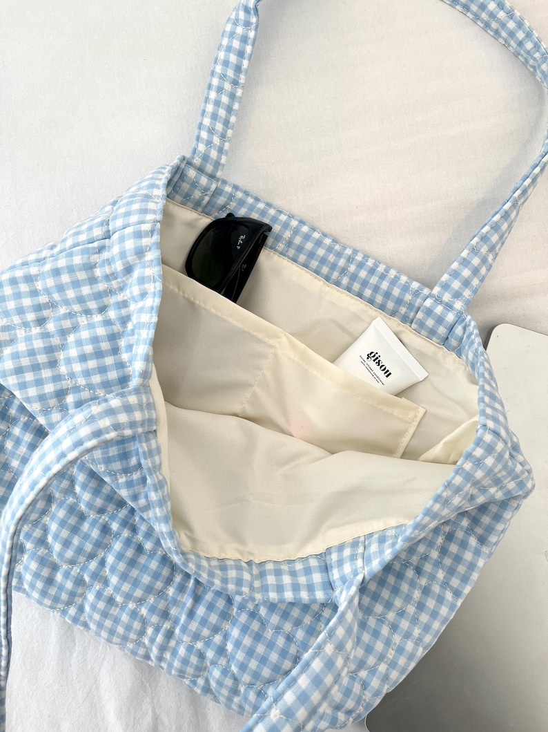 May include: A light blue and white gingham quilted tote bag with two shoulder straps. The interior is lined with cream fabric and features an open pocket containing a pair of black sunglasses and a white tube labeled with the text gison.