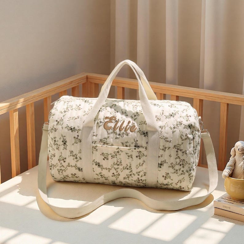 May include: Cream-colored baby bag with a floral pattern and the name "Ellie" embroidered in gold. The bag has a shoulder strap and handles, and is placed on a crib mattress.