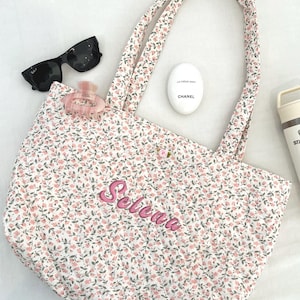 May include: A floral print tote bag with pink embroidered text that reads "Selena". The bag has matching handles and a button closure. Also pictured are black sunglasses, a pink hair clip, and a white Chanel hand cream.