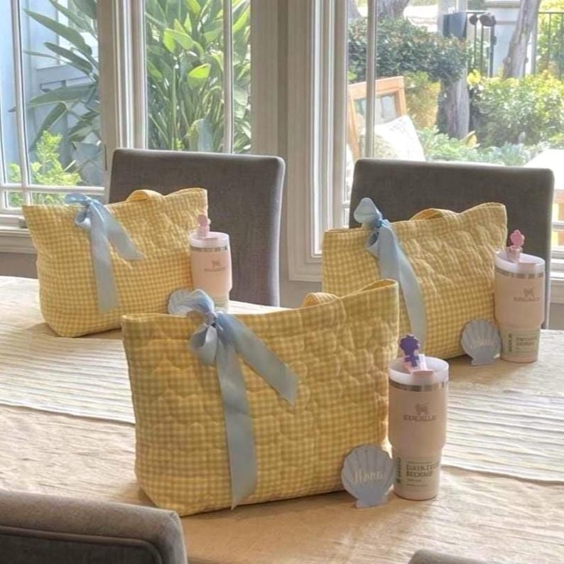 May include: Three yellow and white gingham quilted tote bags, each adorned with a light blue satin ribbon bow. Beside each bag sits a light beige insulated tumbler with a lid and a small decorative seashell-shaped accent piece.