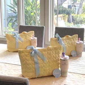 May include: Three quilted tote bags in a yellow and white gingham pattern, each adorned with a light blue ribbon bow. Beside each bag sits a cylindrical tumbler in a light pink shade, accompanied by a small, light blue seashell-shaped tag.