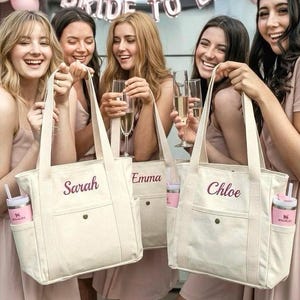 May include: Off-white canvas tote bags with names embroidered in purple. Each bag has a side pocket holding a pink tumbler. The bags are held by women in pink dresses, celebrating a bridal event with champagne.