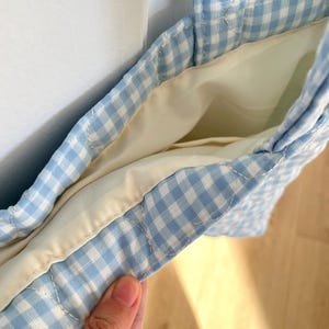 May include: A quilted tote bag featuring a light blue and white gingham check pattern. The bag has a solid cream-colored interior lining and two matching quilted fabric handles. The construction shows visible stitching along the seams and edges.