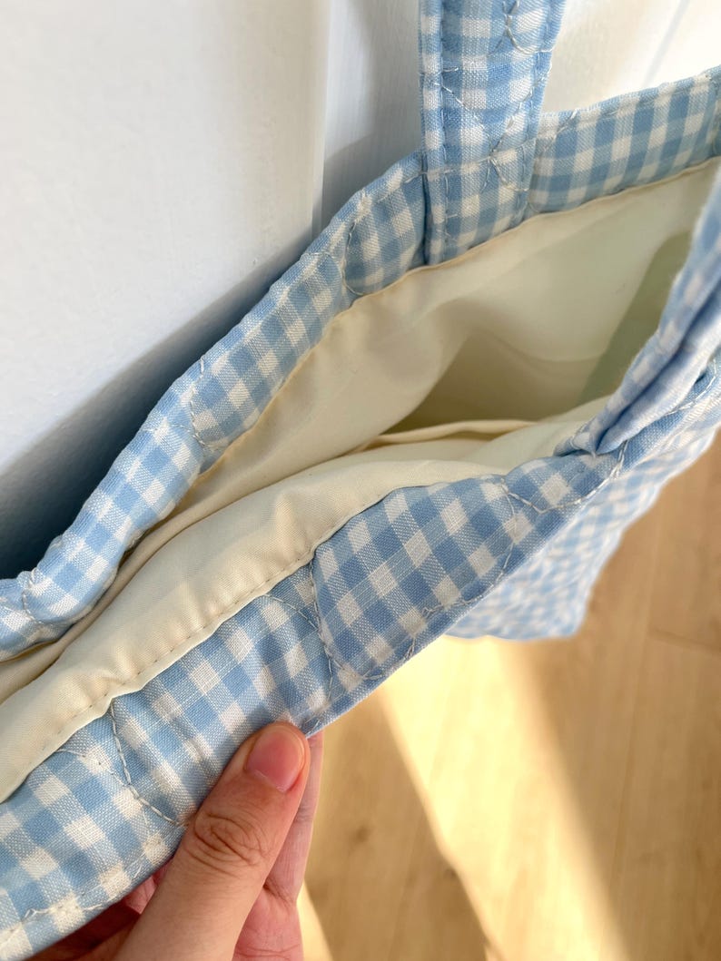 May include: A quilted tote bag featuring a light blue and white gingham check pattern. The bag has a solid cream-colored interior lining and two matching quilted fabric handles. The construction shows visible stitching along the seams and edges.