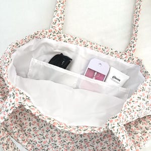 May include: A floral print tote bag with a white interior. The bag features a pattern of small pink flowers and green leaves on a white background. Inside, there are pockets holding a black item, a pink item, and a white tube with the word "gison".
