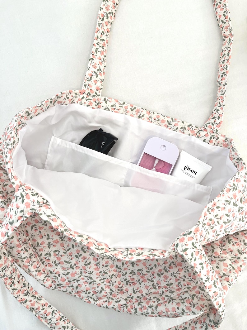 May include: A floral print tote bag with a white interior. The bag features a pattern of small pink flowers and green leaves on a white background. Inside, there are pockets holding a black item, a pink item, and a white tube with the word "gison".