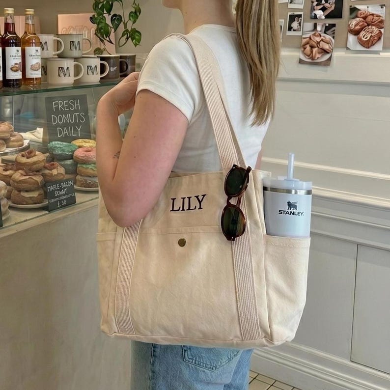 May include: A cream-colored tote bag with the name "LILY" embroidered on the front. The bag has a shoulder strap and a side pocket holding a white tumbler with a straw and sunglasses. The bag is carried by a person in a white t-shirt.