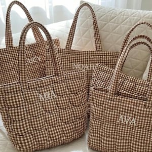 May include: A collection of four quilted tote bags featuring a brown and white gingham check pattern. Each bag has two top handles and is embroidered with a unique name in white thread, specifically the names LUCY, NOAH, MIA, and AVA.