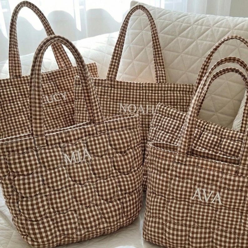 May include: A collection of four quilted tote bags featuring a brown and white gingham check pattern. Each bag has two top handles and is embroidered with a unique name in white thread, specifically the names LUCY, NOAH, MIA, and AVA.