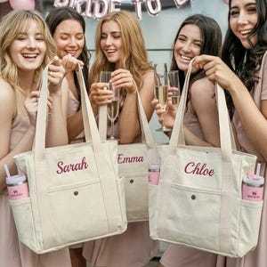 Op de afbeelding: Off-white canvas tote tassen met de namen "Sarah", "Emma" en "Chloe" geborduurd in bordeauxrood. Elke tas heeft een voorvak en een zijvak met een roze beker. Op de achtergrond een "Bride to Be" banner en roze ballonnen.