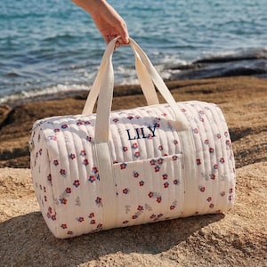 May include: A cream-colored quilted duffel bag with a floral pattern of red and blue flowers. The bag has cream-colored handles and a pocket on the front. The name "LILY" is embroidered in navy blue on the side of the bag.