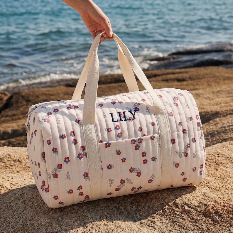 May include: A cream-colored quilted duffel bag with a floral pattern of red and blue flowers. The bag has cream-colored handles and a pocket on the front. The name "LILY" is embroidered in navy blue on the side of the bag.