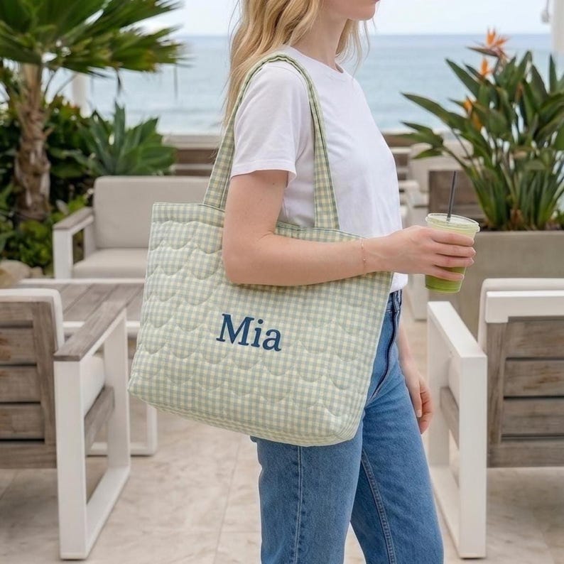 May include: A light green and white gingham quilted tote bag featuring the name Mia embroidered in dark blue thread on the front. The bag has two shoulder straps and a rectangular shape with a soft, padded texture throughout the fabric.