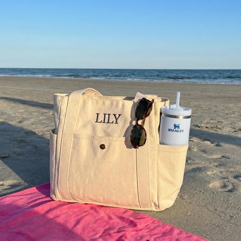 May include: A beige canvas tote bag with the name "LILY" embroidered on the front. The bag has side pockets and a button closure. Sunglasses and a white Stanley tumbler with a straw are in the pockets. The bag is on a sandy beach.