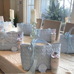 May include: Several quilted tote bags featuring a light blue and white gingham pattern, each adorned with a light blue ribbon bow. Accompanying the bags are light pink insulated tumblers with straws and small shell-shaped name tags on the table.
