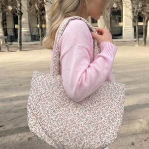 May include: A floral patterned tote bag in pink and green. The bag has a rounded shape and a matching strap. The background is a blurred outdoor scene. The bag appears to be quilted.