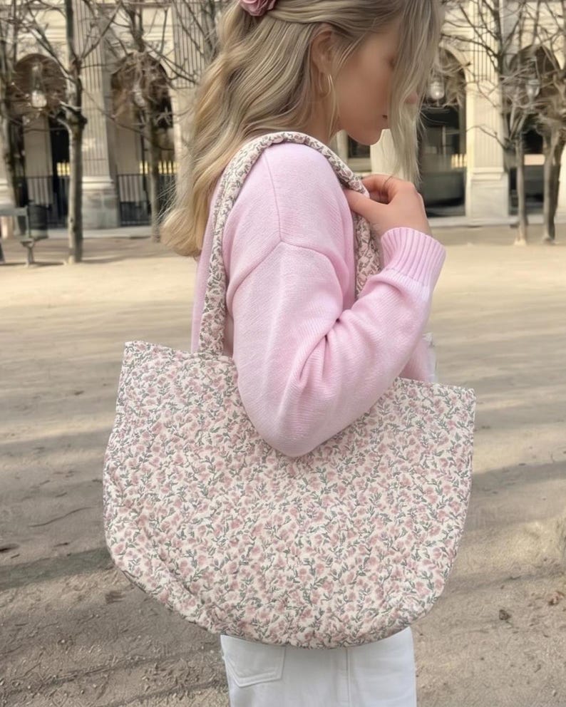 May include: A floral patterned tote bag in pink and green. The bag has a rounded shape and a matching strap. The background is a blurred outdoor scene. The bag appears to be quilted.