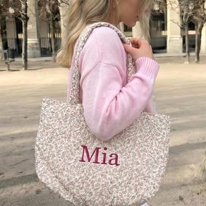 May include: A quilted tote bag with a floral pattern in pink and green. The word "Mia" is embroidered in burgundy on the front. The bag has matching floral straps. The bag is being carried over the shoulder.