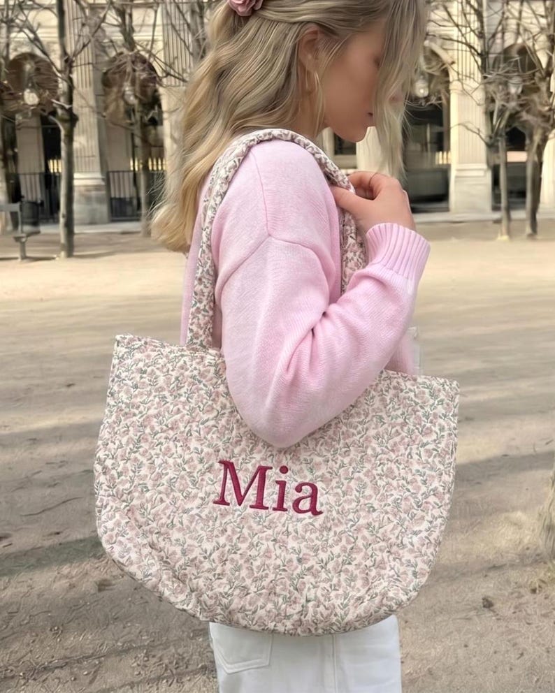 May include: A quilted tote bag with a floral pattern in pink and green. The word "Mia" is embroidered in burgundy on the front. The bag has matching floral straps. The bag is being carried over the shoulder.