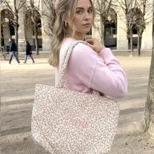 May include: A pink floral tote bag embroidered with the name "Tara" in pink script. The bag has matching floral straps and is carried over the shoulder. The background shows a person wearing a pink sweater.