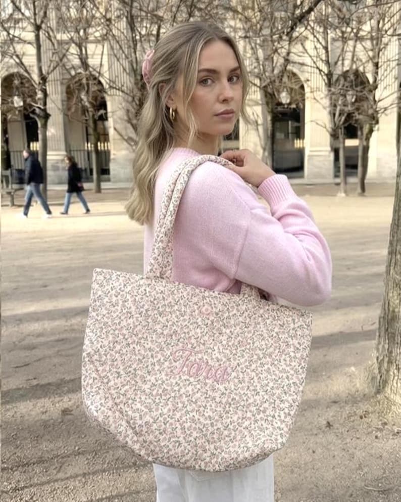May include: A pink floral tote bag embroidered with the name "Tara" in pink script. The bag has matching floral straps and is carried over the shoulder. The background shows a person wearing a pink sweater.