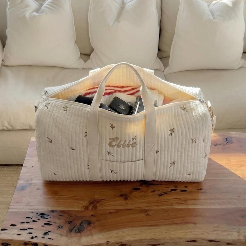 May include: Cream-colored quilted duffel bag with a top handle and embroidered floral accents. The bag is open, revealing contents including a red and white striped towel and other items. The name "Ellie" is embroidered on the front.