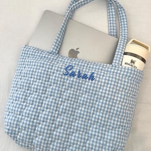 May include: A light blue and white gingham quilted tote bag with two matching fabric handles. The front of the bag features the name Sarah embroidered in blue script. A silver laptop and a cream-colored Stanley brand tumbler are inside.