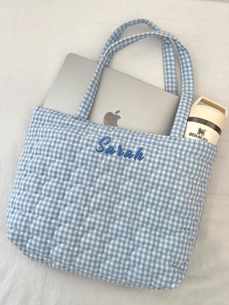 May include: A light blue and white gingham quilted tote bag with two matching fabric handles. The front of the bag features the name Sarah embroidered in blue script. A silver laptop and a cream-colored Stanley brand tumbler are inside.