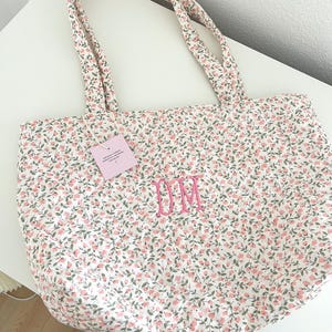 May include: A floral tote bag featuring a pattern of pink flowers and green leaves on a white base. The bag has matching handles and embroidered pink initials "DM". A small tag is attached.