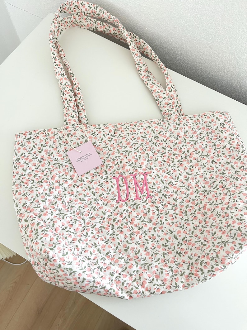 May include: A floral tote bag featuring a pattern of pink flowers and green leaves on a white base. The bag has matching handles and embroidered pink initials "DM". A small tag is attached.