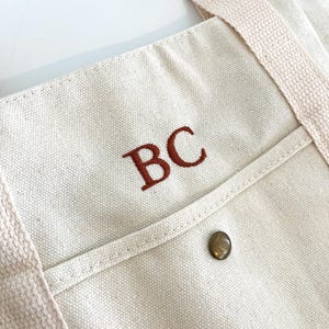 May include: Cream-colored canvas tote bag with a front pocket and brown embroidered initials "BC". The bag has matching cream-colored straps and a metal button closure on the pocket. Sunglasses are visible in the background.