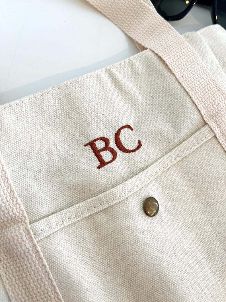 May include: Cream-colored canvas tote bag with a front pocket and brown embroidered initials "BC". The bag has matching cream-colored straps and a metal button closure on the pocket. Sunglasses are visible in the background.