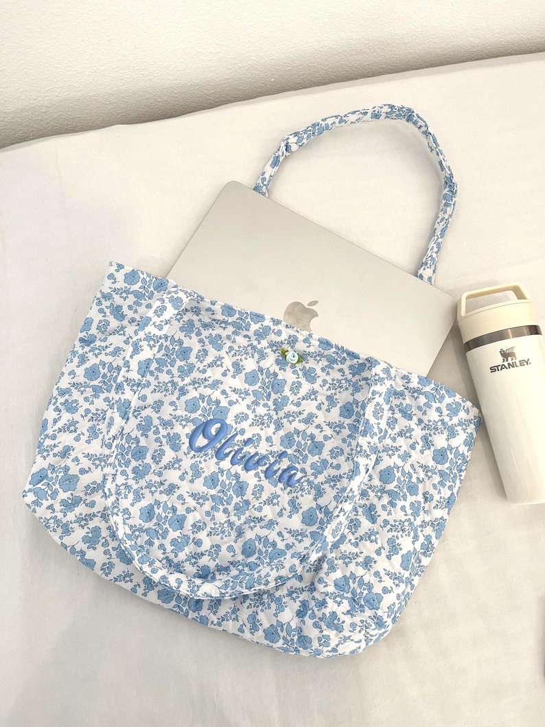 May include: A white and light blue floral tote bag with matching handles. The bag holds a laptop and a white and beige insulated water bottle. The name "Olivia" is embroidered on the front of the bag.