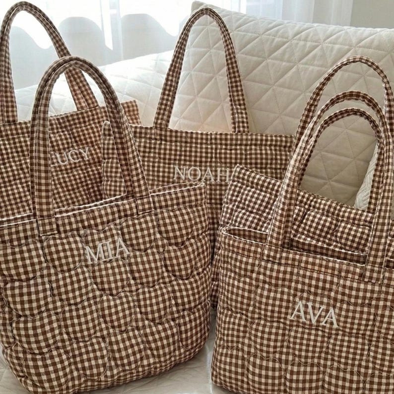 May include: A collection of four quilted tote bags featuring a brown and white gingham check pattern. Each bag has two fabric handles and is personalized with white embroidered names including Lucy, Noah, Mia, and Ava on the front panels.