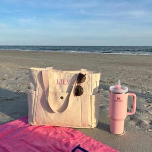 May include: A beach scene featuring a light beige tote bag with the name "LILY" embroidered in pink, sunglasses, and a pink Stanley tumbler with a straw. A pink beach towel is in the foreground, with the ocean in the background.