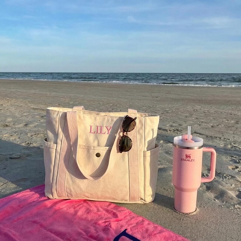 May include: A beach scene featuring a light beige tote bag with the name "LILY" embroidered in pink, sunglasses, and a pink Stanley tumbler with a straw. A pink beach towel is in the foreground, with the ocean in the background.
