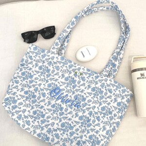 May include: A white tote bag featuring a blue floral design and matching handles. The name "Olivia" is embroidered in blue script. Black sunglasses, a white oval cosmetic container, and a white Stanley flask are also present.