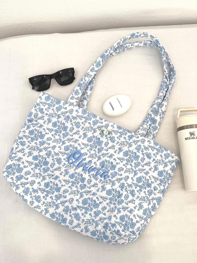 May include: A white tote bag featuring a blue floral design and matching handles. The name "Olivia" is embroidered in blue script. Black sunglasses, a white oval cosmetic container, and a white Stanley flask are also present.