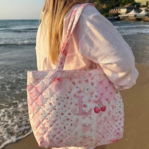 May include: A pink quilted tote bag with a floral and bow design. The bag displays the letter "L" and cherries. The bag has a diamond pattern and pink straps.