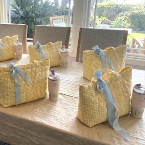 May include: Four quilted yellow and white gingham tote bags, each tied with a light blue ribbon, arranged on a table alongside four matching light pink insulated tumblers with lids, creating a coordinated set of gift bags and drinkware.