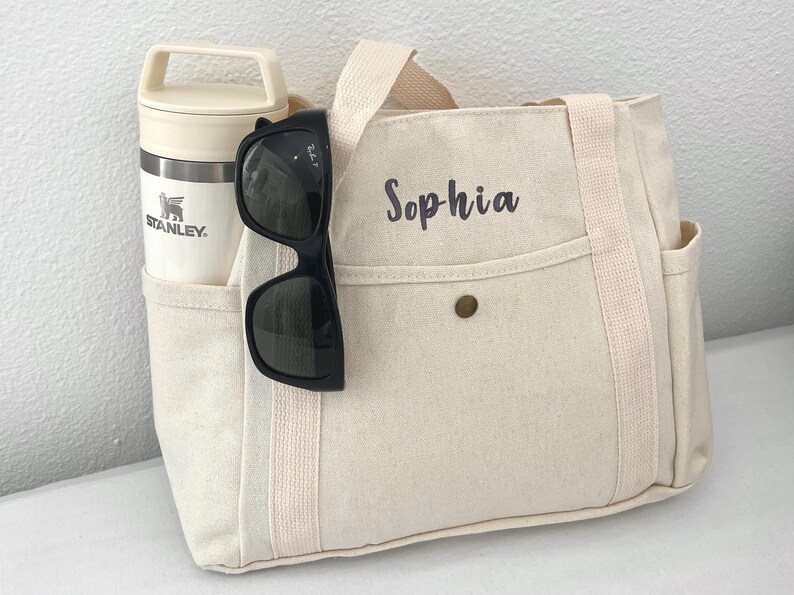 May include: A cream-colored canvas tote bag with the name "Sophia" printed on the front. The bag has side pockets and a beige handle. A black pair of sunglasses hangs on the front, and a cream-colored Stanley thermos is in the side pocket.