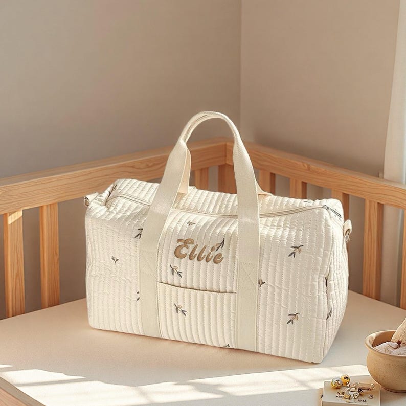 May include: Cream-colored quilted baby bag with the name "Ellie" embroidered on the front. The bag has a rectangular shape with a top handle and a front pocket. Small leaf patterns are embroidered across the bag.