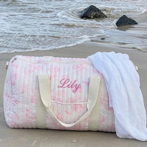 May include: A pink floral quilted duffel bag with the name "Lily" embroidered on the front. The bag has cream-colored handles and a white towel draped over it. The bag is on a sandy beach near the ocean.