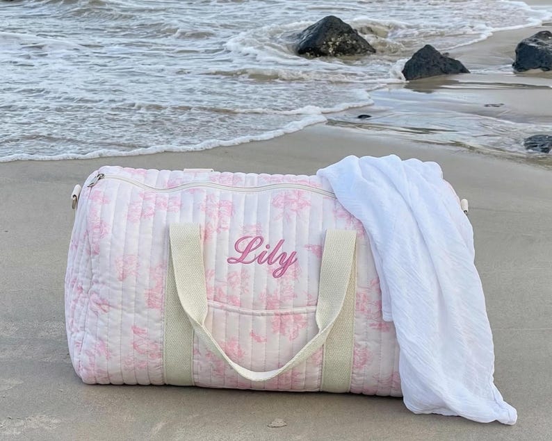 May include: A pink floral quilted duffel bag with the name "Lily" embroidered on the front. The bag has cream-colored handles and a white towel draped over it. The bag is on a sandy beach near the ocean.