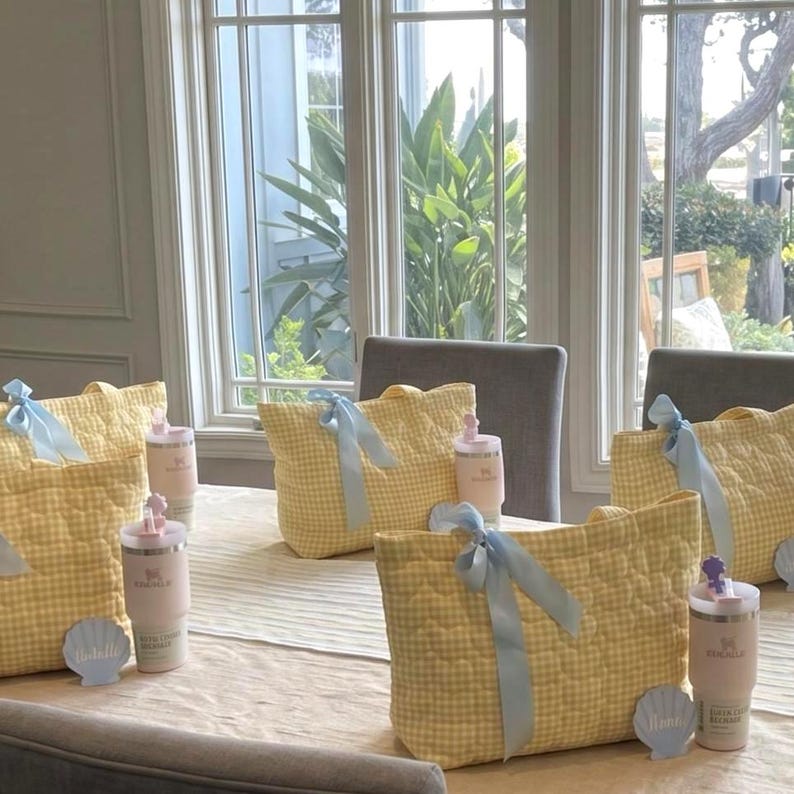 May include: Three quilted tote bags in a yellow and white gingham pattern, each tied with a light blue ribbon. Beside each bag sits a light pink insulated tumbler with a straw and a small, light blue seashell-shaped name tag.