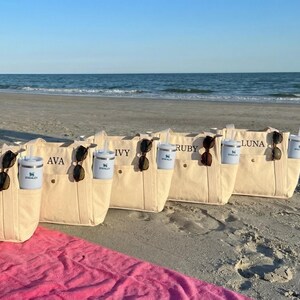 May include: Beige canvas tote bags with personalized names, sunglasses, and insulated tumblers with straws. The bags are on a sandy beach with the ocean in the background. A pink towel is in the foreground.