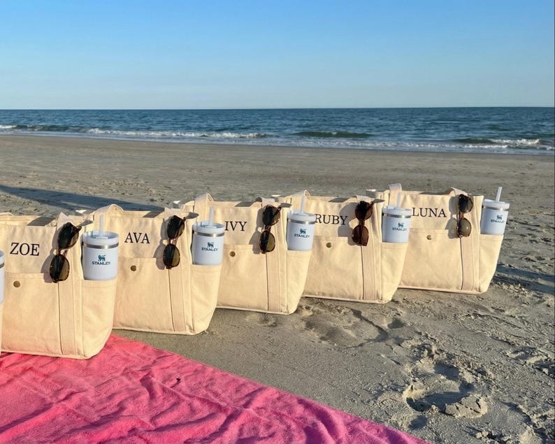 May include: Beige canvas tote bags with personalized names, sunglasses, and insulated tumblers with straws. The bags are on a sandy beach with the ocean in the background. A pink towel is in the foreground.
