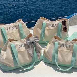 May include: Four cream-colored canvas tote bags with mint green accents and handles. Each bag features a personalized name embroidered in grey: LUNA, SARA, MIA, and RUBY. One bag includes a grey patterned bow, and another has sunglasses attached.